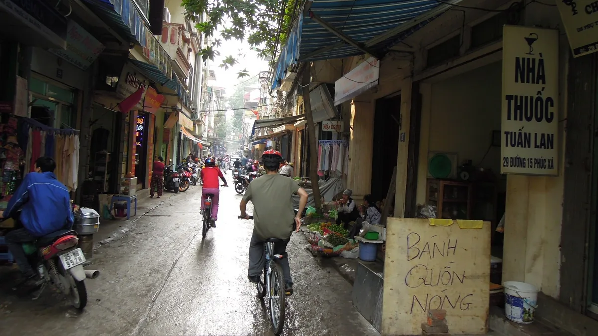 Cyclists and motorbikes navigate a narrow Hanoi alley lined with shops, awnings and a pharmacy sign.