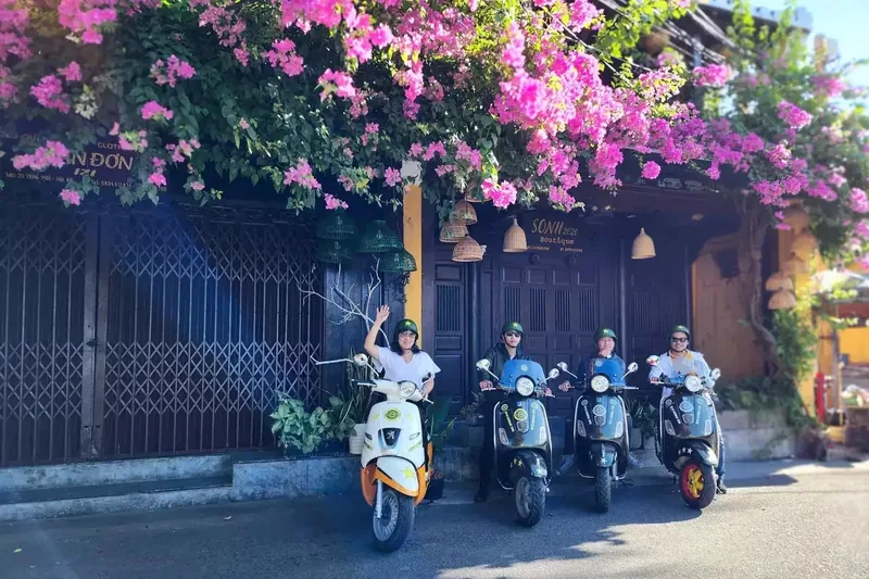 Riders on scooters pose beneath a bougainvillea-covered shopfront on a quiet Hoi An street.