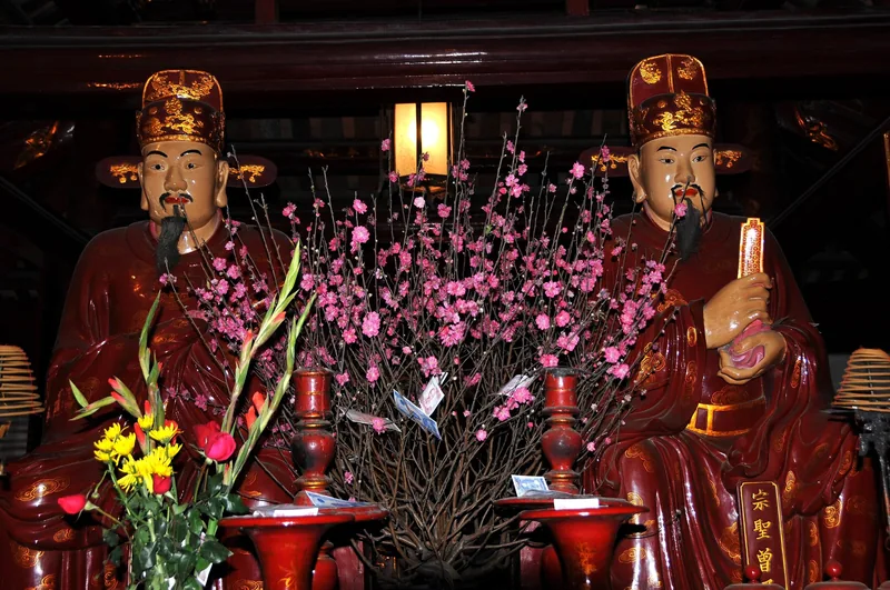 Two red-robed statues flank a pink peach blossom branch inside a temple in Hanoi.