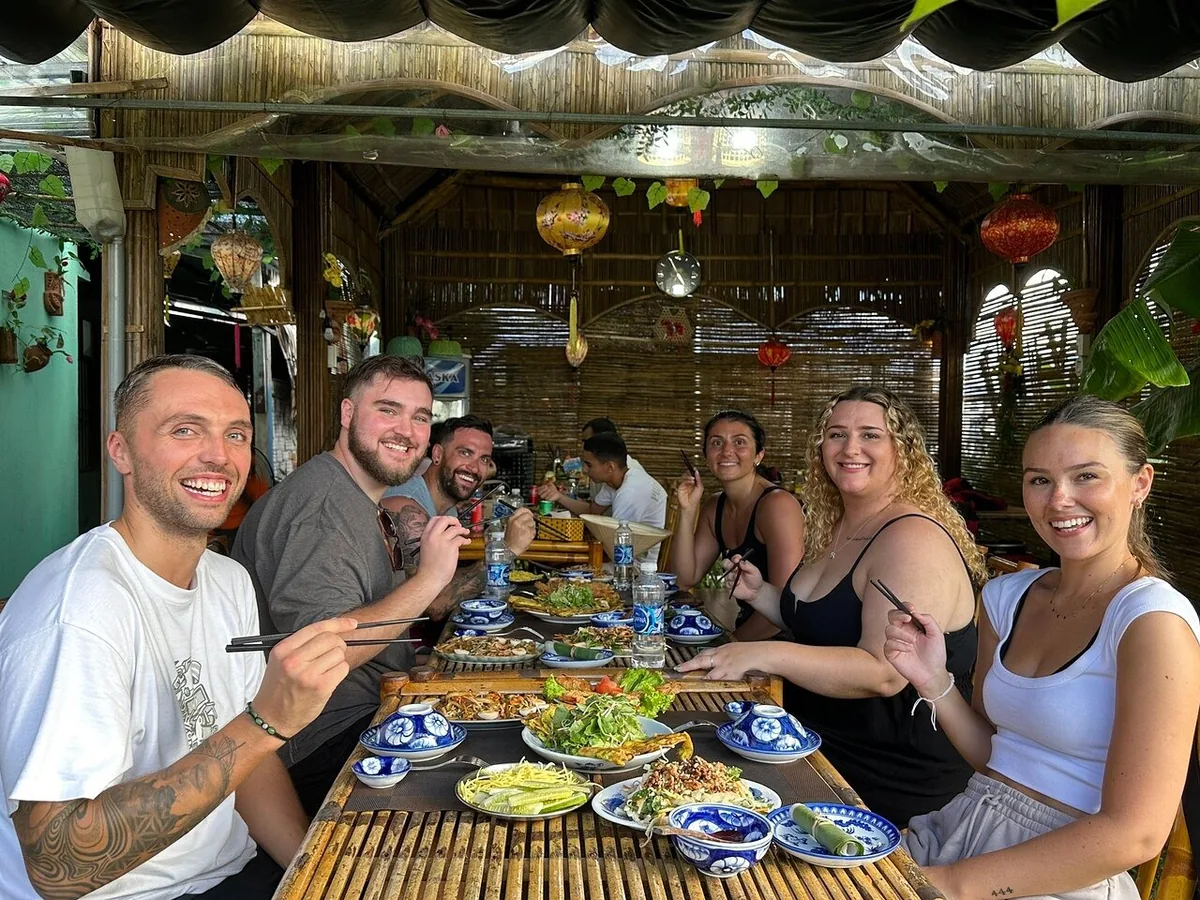 A group of tourists smiles with chopsticks over a table of local dishes at a bamboo restaurant in Tra Que village.