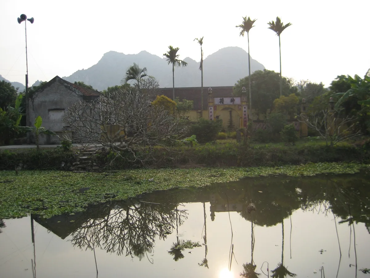 A tranquil pond reflects tall palms and limestone mountains beside a rural temple in northern Vietnam.