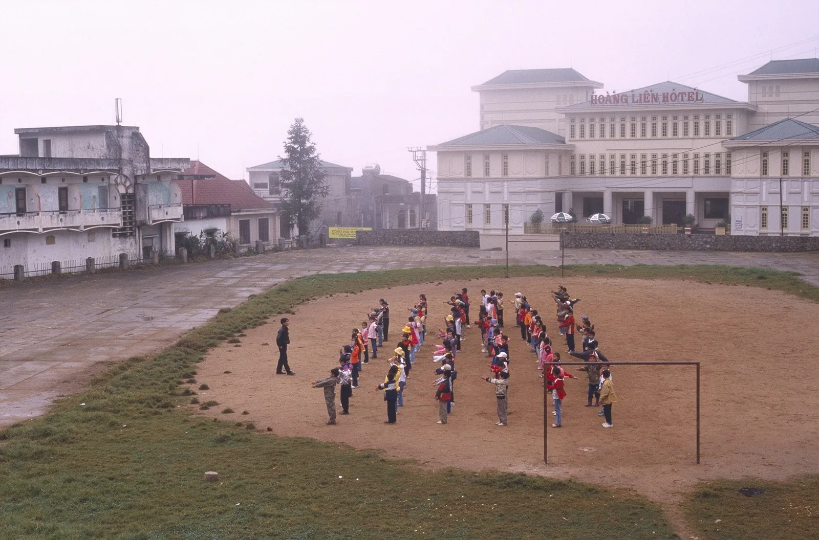 Children line up for morning exercises on a dirt square in Sapa, with Hoang Lien Hotel in the misty background.