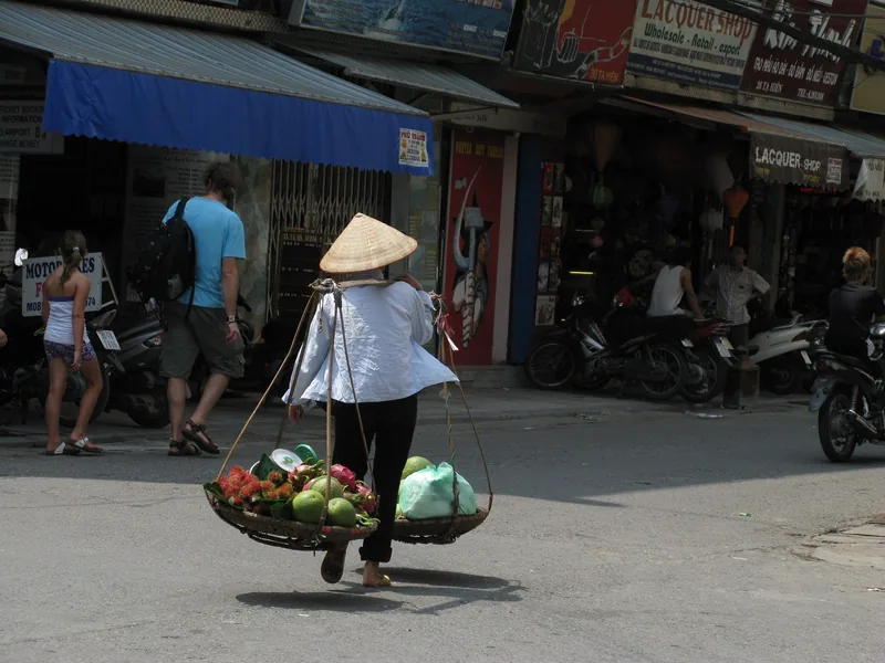 A conical-hatted street vendor carries fruit baskets on a shoulder pole through a busy Hanoi street lined with shops.
