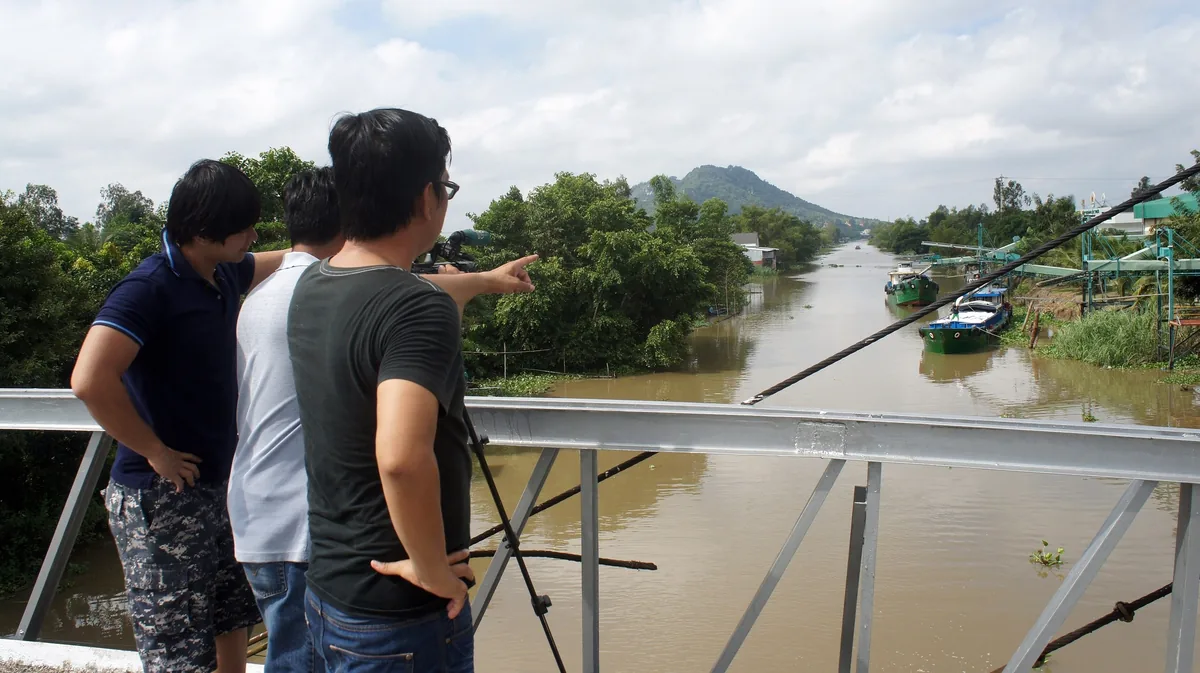 Three men on a bridge point toward a brown river with boats and a distant mountain near Ho Chi Minh City