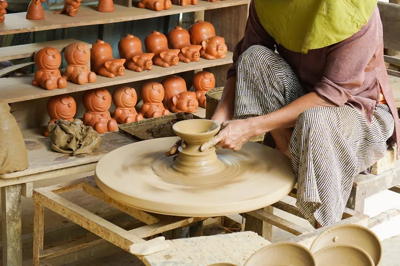 An artisan shapes clay on a spinning wheel beside shelves of terracotta figurines at a Hoi An craft village.