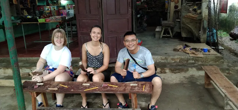 Three tourists sit on a wooden bench trying woodcarving tools at a traditional craft village in Hoi An.