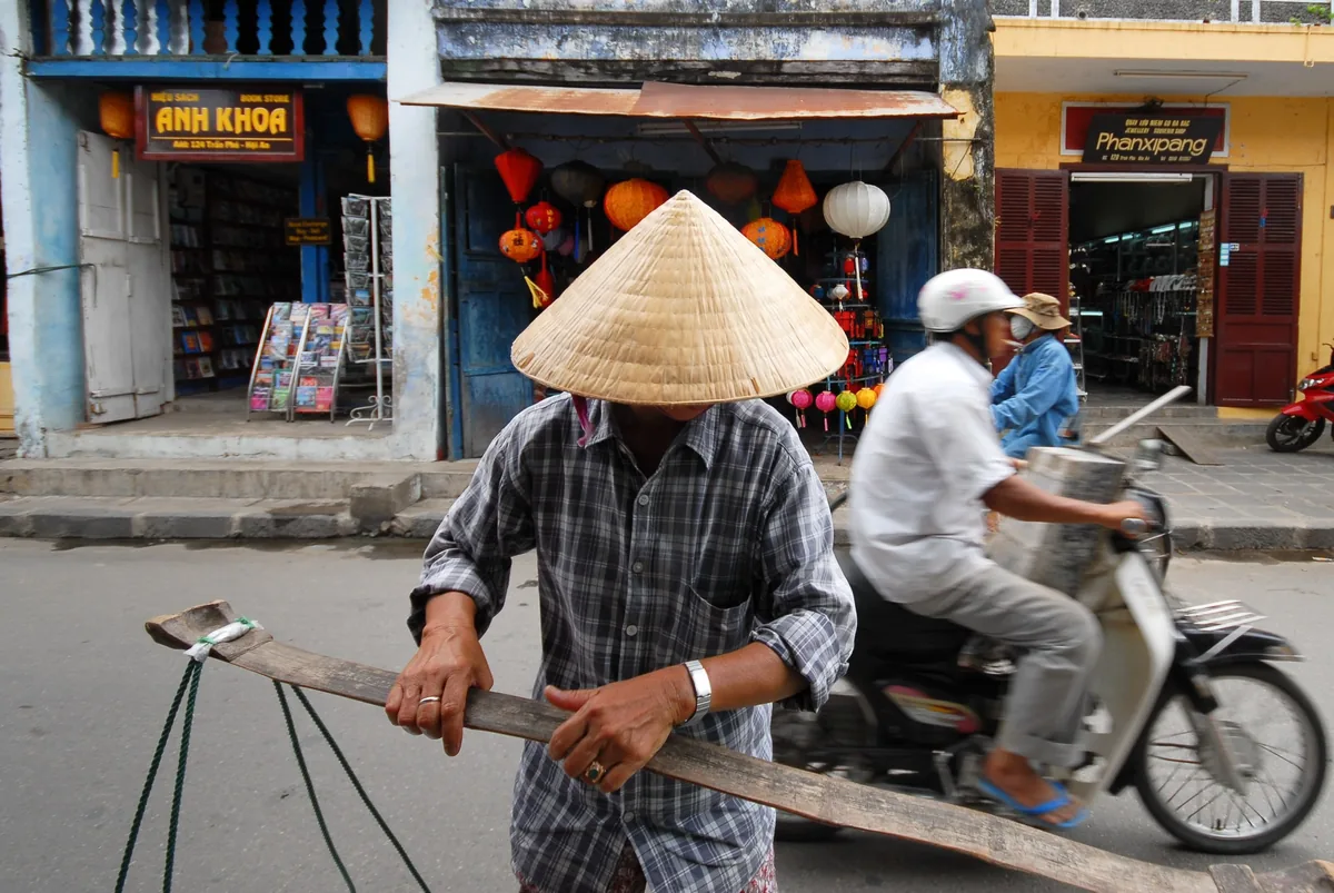 A conical hat vendor carries a shoulder pole past colorful shops as a motorbike passes on a Hoi An street.