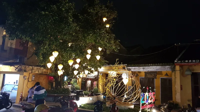 Yellow lanterns glow in a tree above a 2025 New Year decoration on a quiet street in Hoi An's old town at night.