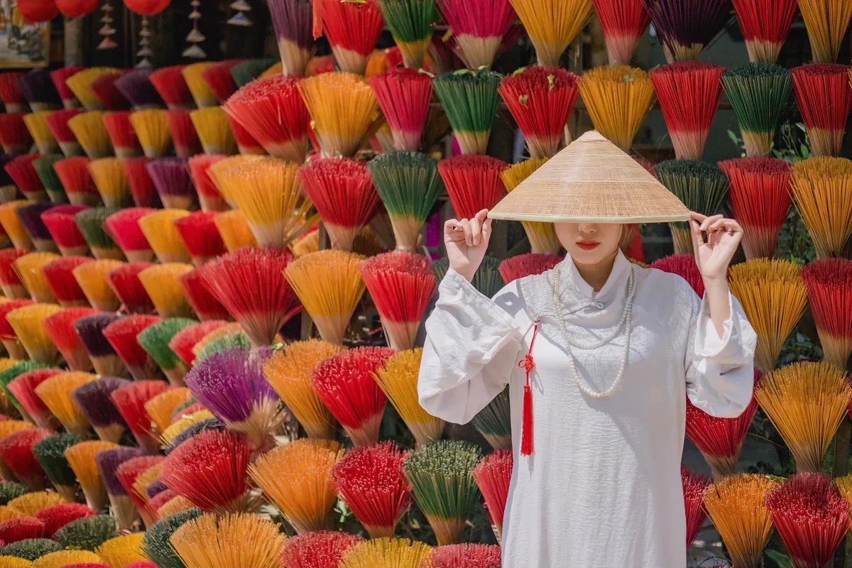 A woman in a white ao dai and conical hat poses among colorful incense sticks in Hue.