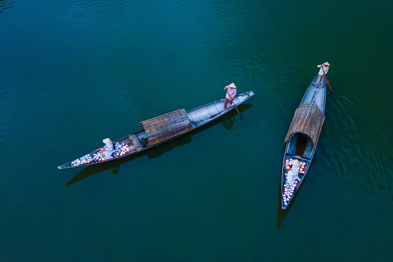 Aerial view of two traditional wooden boats with conical-hat fishermen crossing calm green river water in Vietnam.