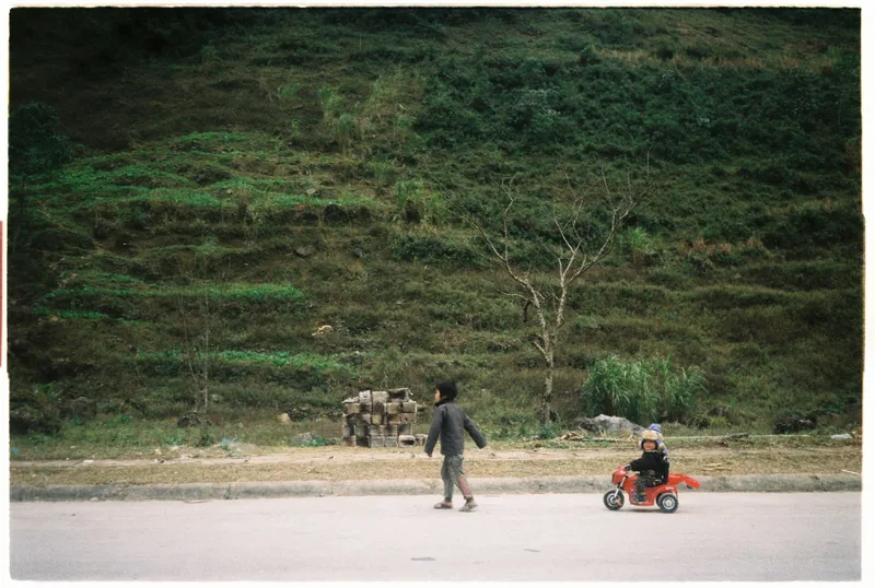Children play on a quiet road with a toy motorbike below a terraced green hillside in Ha Giang, Vietnam.