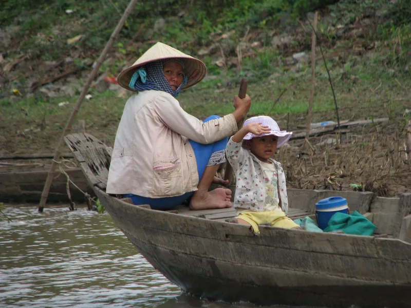 A woman in a conical hat and a small child wave from a wooden boat on a Mekong Delta river.
