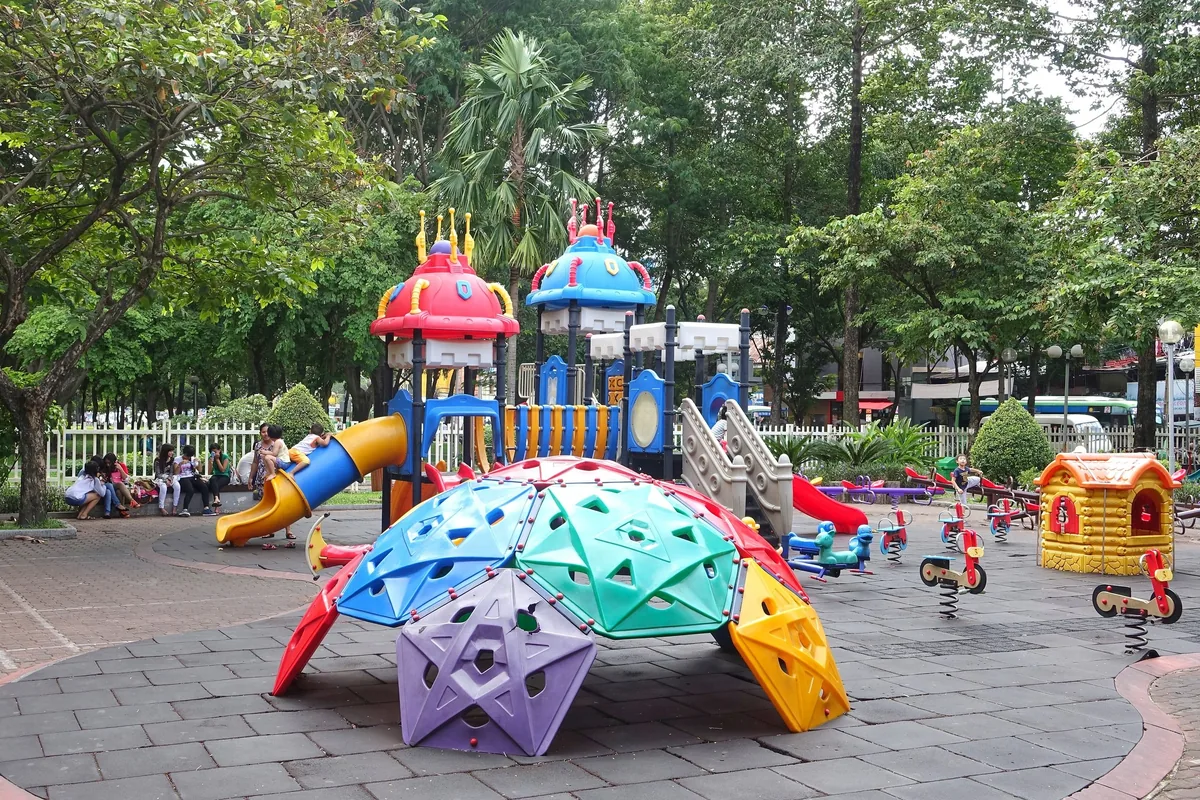 Colorful playground equipment sits in a shaded park in Ho Chi Minh City with parents watching children nearby.
