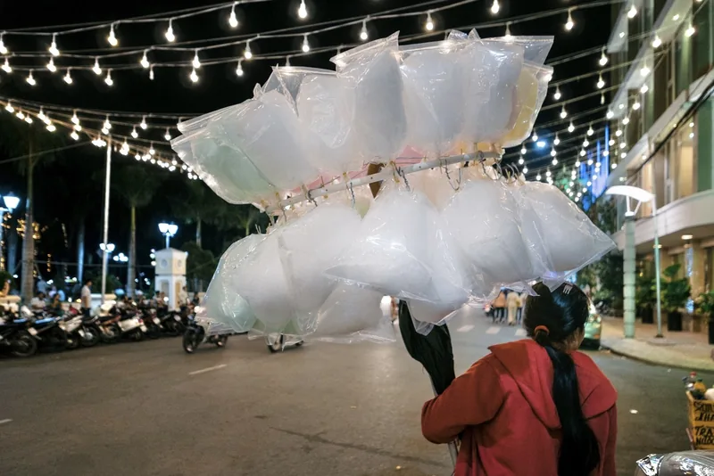 A street vendor carries bags of cotton candy under string lights on Hai Ba Trung Street in Can Tho at night.