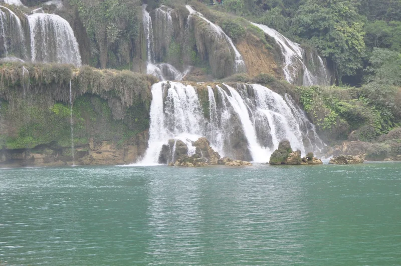 Multiple tiers of cascading water flow over limestone cliffs into a turquoise pool at Ban Gioc Waterfall.