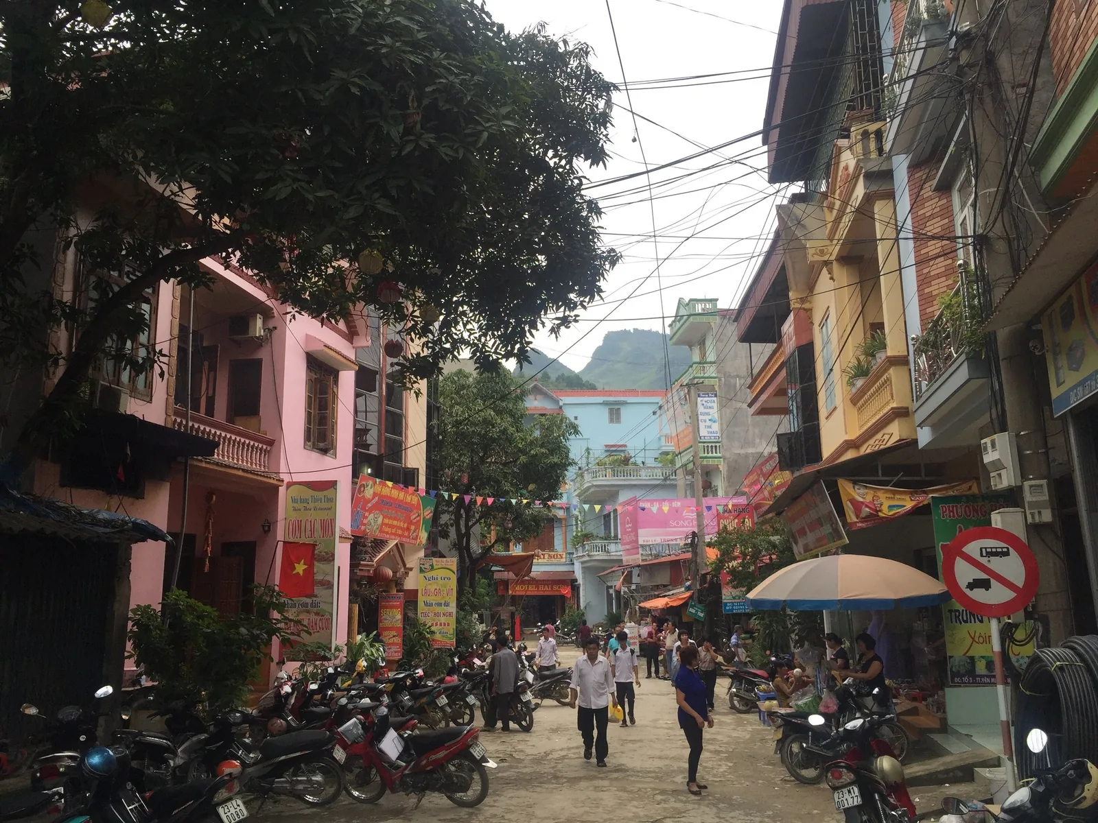 A narrow street in Ha Giang lined with colorful houses, parked motorbikes and a mountain backdrop.