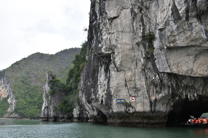A limestone karst tunnel with a sign reading Hang Luon, small boats carrying tourists in life vests entering the cave in Ha.