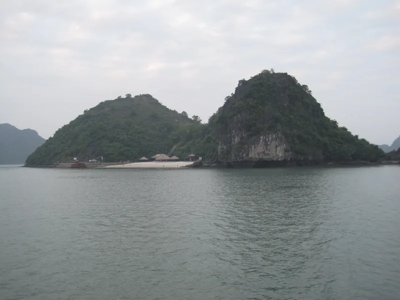 A small sandy beach with thatched huts sits between two limestone islands in Ha Long Bay.