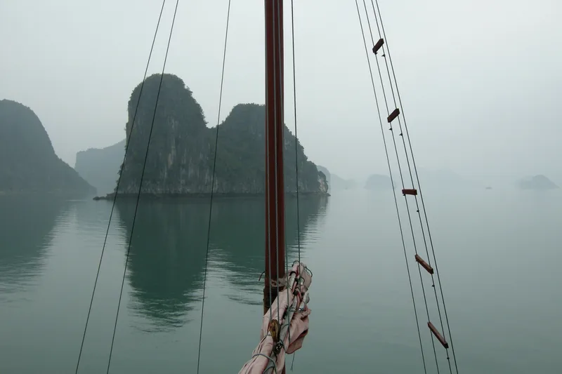 A wooden mast and rigging frame a misty view of limestone karsts rising from Ha Long Bay's calm water.