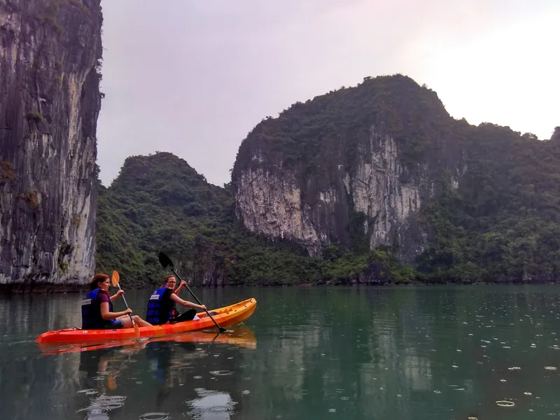 Two women paddle an orange kayak past towering limestone karsts in Ha Long Bay.