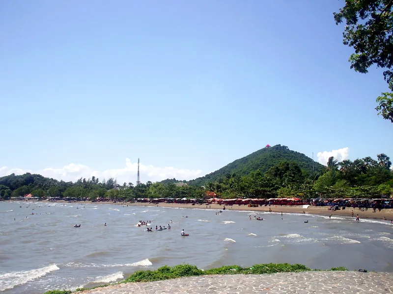 Swimmers and beach umbrellas line the shore at Mui Nai beach in Ha Tien with a forested hill behind.