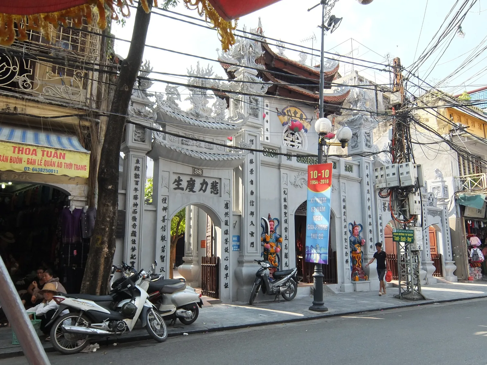 An ornate stone temple gate with carved dragons stands on a busy Hanoi street lined with shops and parked motorbikes.