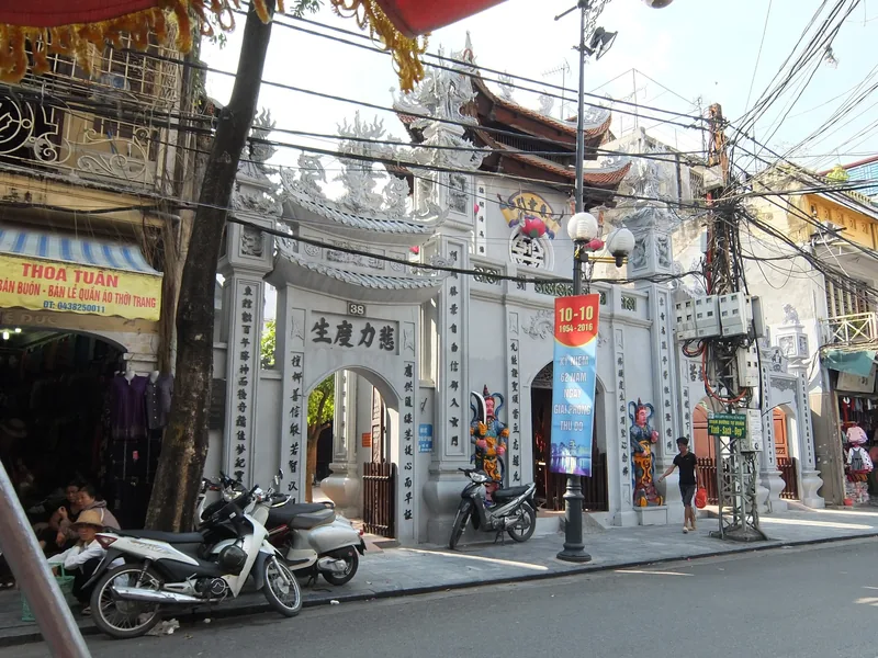 An ornate stone temple gate with carved dragons stands on a busy Hanoi street lined with shops and parked motorbikes.