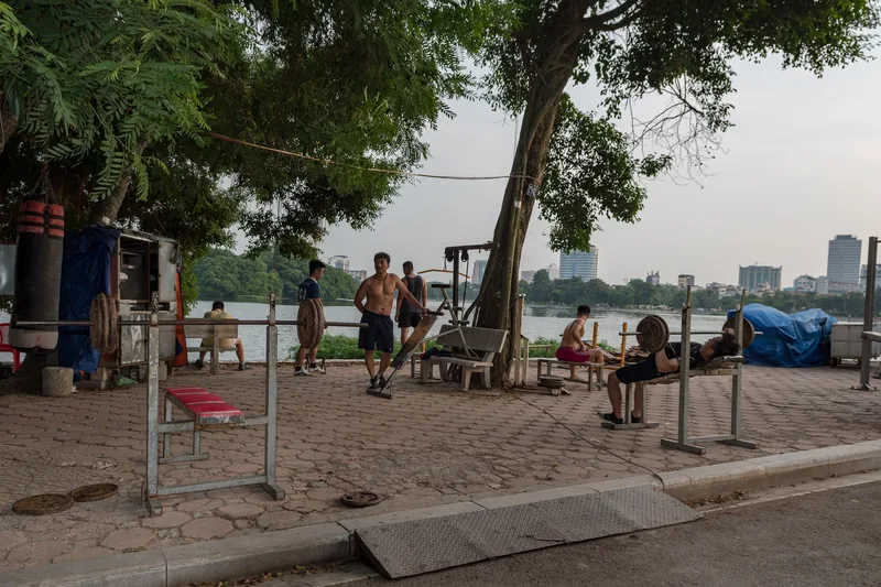 Locals use outdoor exercise equipment beside a lake, with the Hanoi skyline visible across the water.