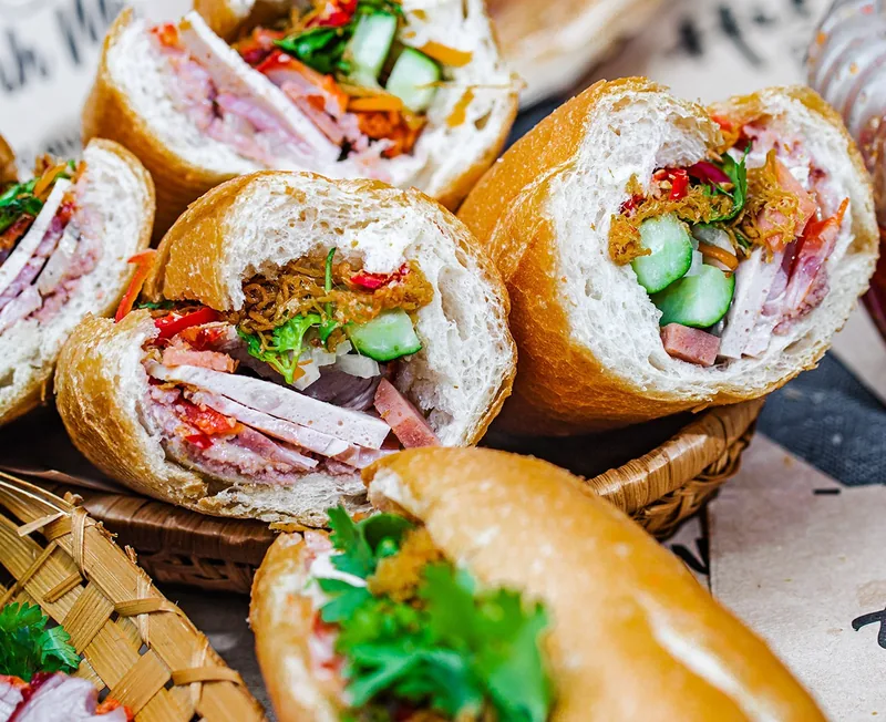 Sliced banh mi sandwiches filled with pork, pate, herbs, and pickled vegetables in a bamboo basket.