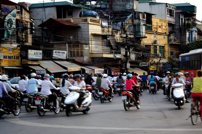 Motorbikes crowd a busy street intersection lined with narrow tube houses in Hanoi's Old Quarter.