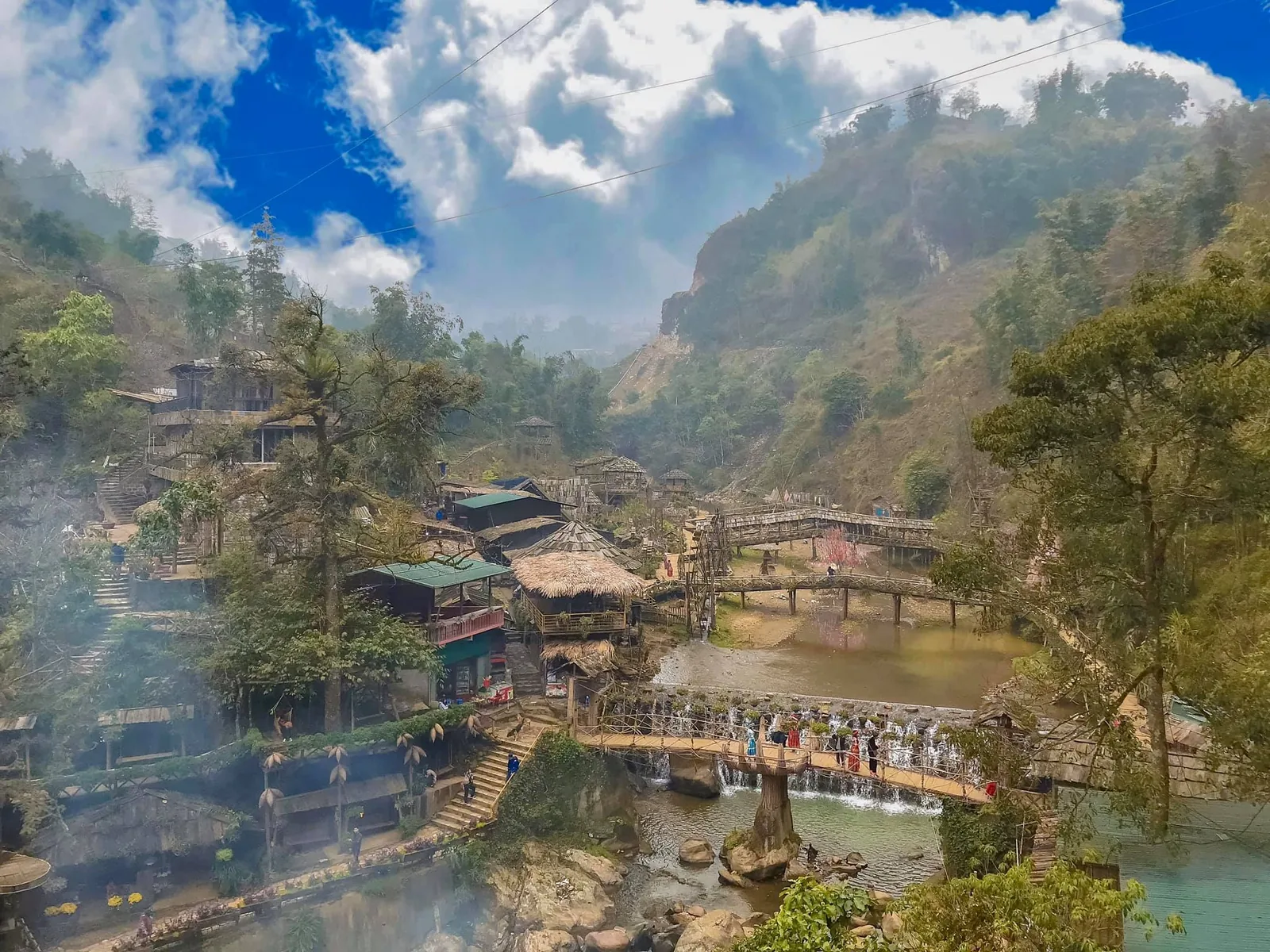 A mountain village near Sapa with thatched huts, small waterfalls and bridges over a stream surrounded by misty peaks.