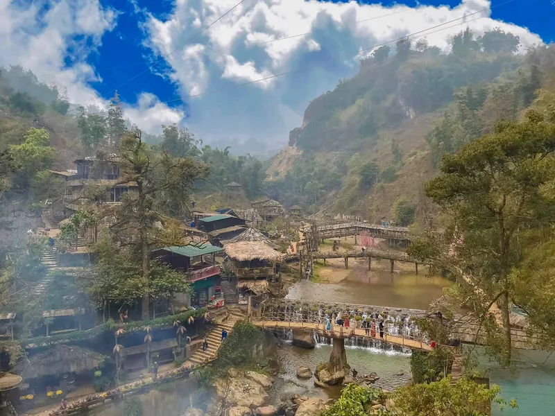 A mountain village near Sapa with thatched huts, small waterfalls and bridges over a stream surrounded by misty peaks.