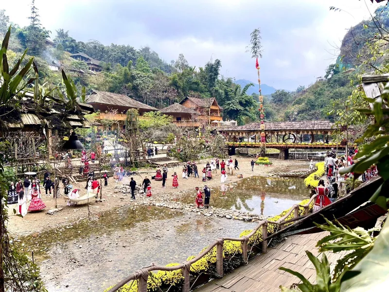 Visitors in colorful outfits gather by a stream lined with yellow flowers in Cat Cat Village, Lao Cai.
