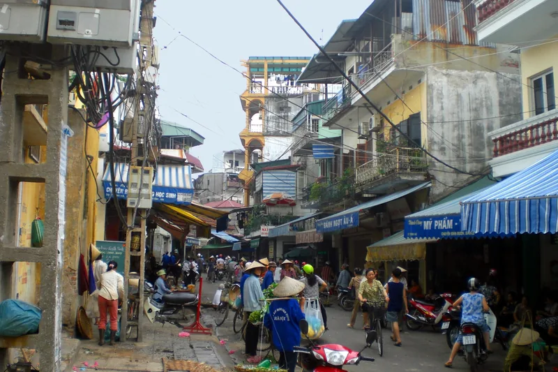 A busy street market in Hanoi's Old Quarter with vendors, motorbikes and tangled overhead wires between narrow houses.