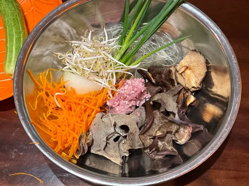 A metal bowl holds ingredients for Vietnamese spring rolls, including shredded carrot, mushrooms, sprouts, and minced meat.