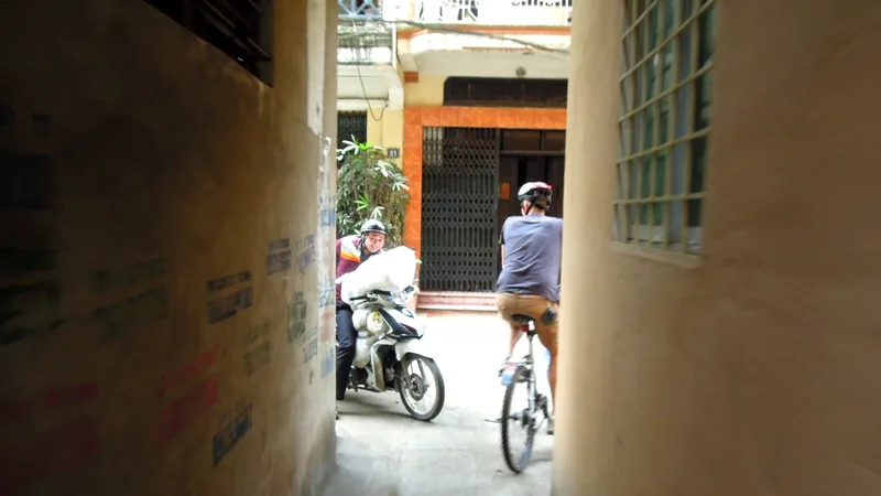 A cyclist rides through a narrow residential alley past a parked motorbike in Hanoi.