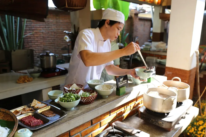 A chef in white uniform prepares fresh pho at an outdoor cooking station with herbs and broth pots in Hoi An.