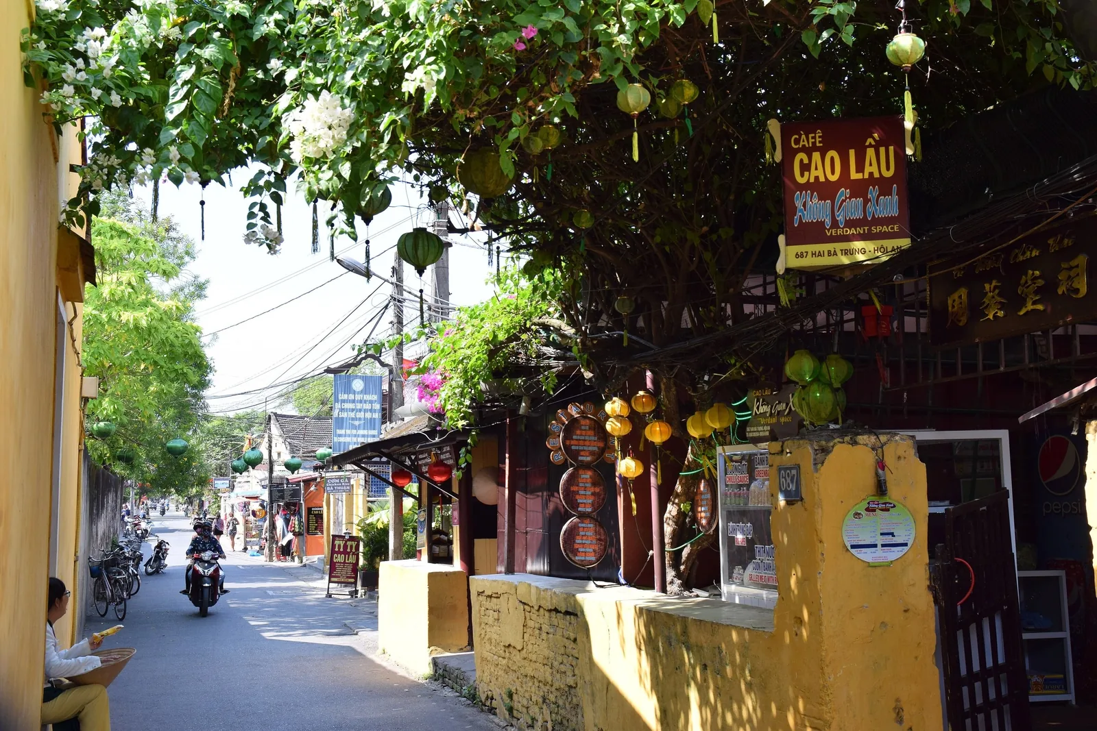 A tree-lined street in Hoi An with yellow buildings, hanging lanterns and a cafe sign along the sidewalk.