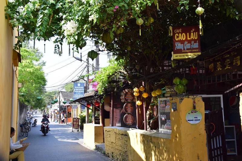 A tree-lined street in Hoi An with yellow buildings, hanging lanterns and a cafe sign along the sidewalk.