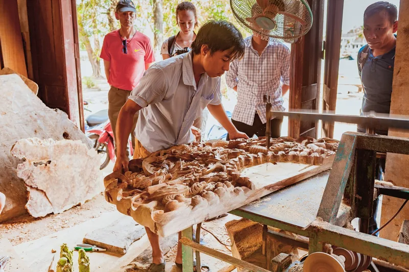 A craftsman shapes an ornately carved wooden panel while visitors watch at Kim Bong Carpentry Village in Hoi An.