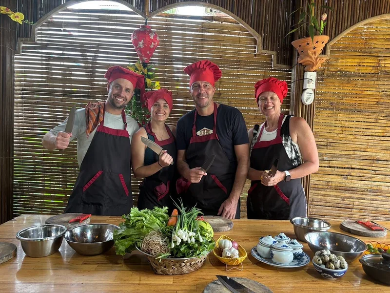 Tourists in red chef hats and aprons pose with knives beside fresh herbs at a Tra Que cooking class