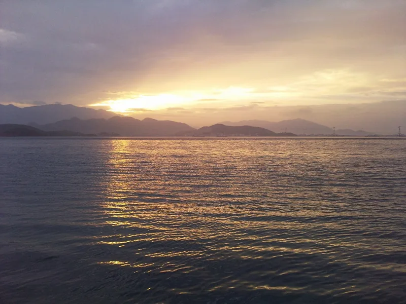 Golden sunset light spreads across the calm waters of Nha Trang Bay with mountains on the horizon.