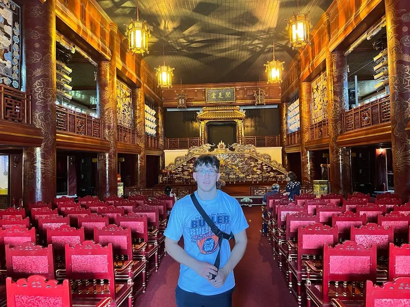 A visitor stands amid rows of red lacquered chairs inside an ornate golden throne hall in Hue.
