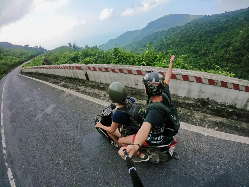 Two riders on a motorbike raise a hand while cruising a winding mountain road on the Hue to Hoi An route.