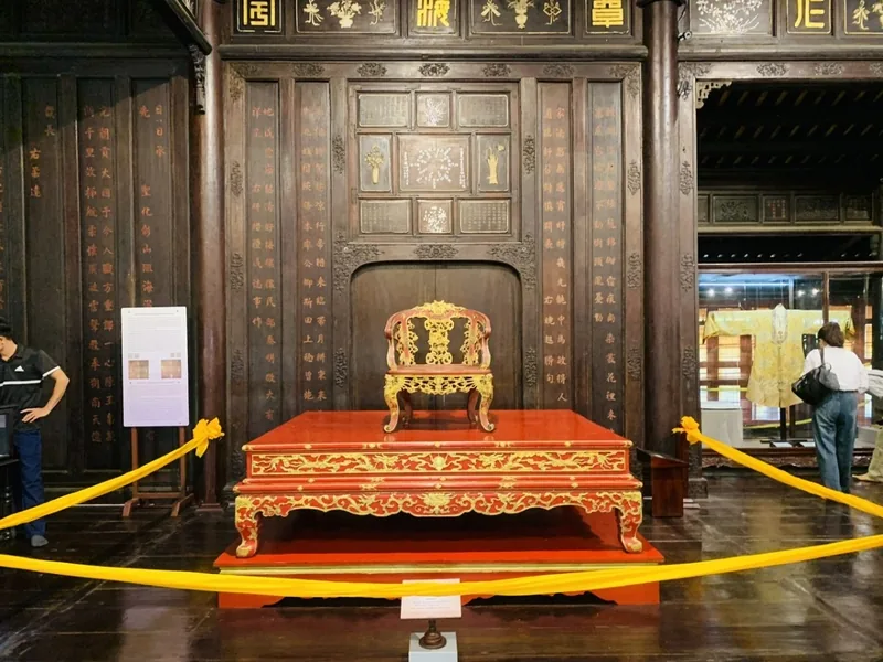 A carved gilded royal throne on a red lacquered platform inside a wooden-paneled hall in Hue.