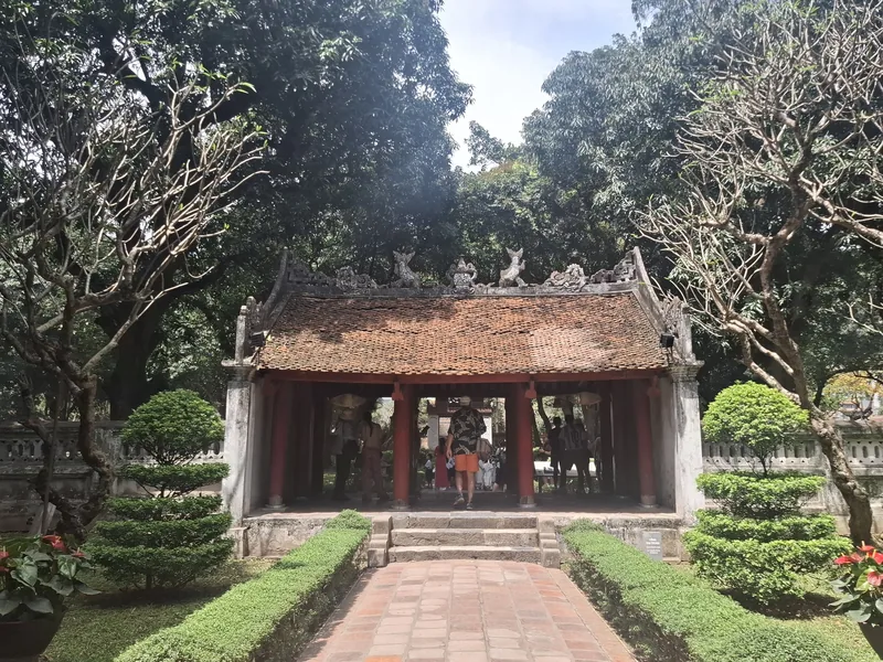 A red-pillared pavilion with a tiled roof stands amid trimmed hedges and trees at a Hanoi temple complex.