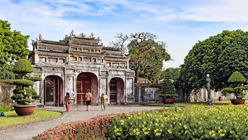 An ornate ancient gate leads visitors into the Hue Imperial Citadel, framed by flower gardens and bonsai trees.