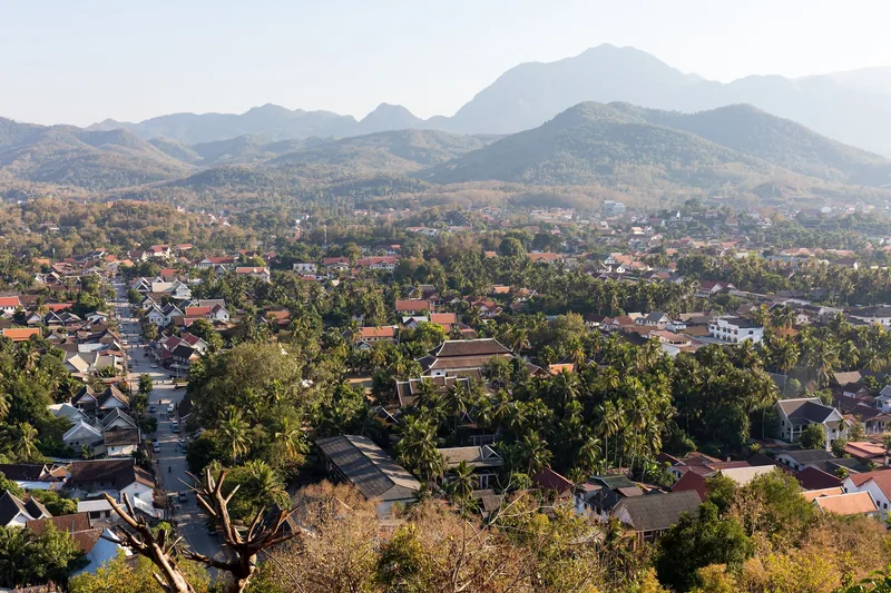 Aerial view of Luang Prabang's tree-lined streets and rooftops surrounded by misty mountains, seen from Phousi Hill.