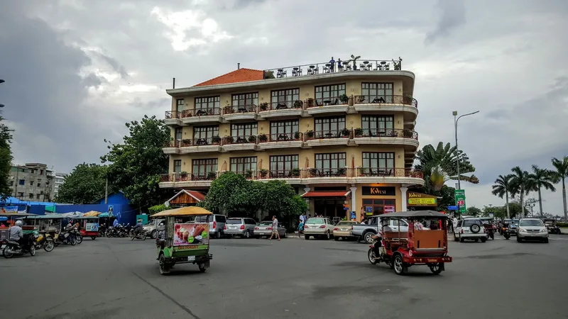 Tuk-tuks pass a rounded corner hotel building along Preah Sisowath Quay in Phnom Penh under a cloudy sky.