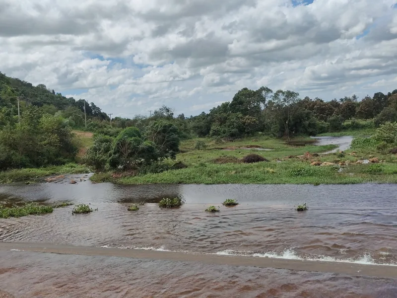 A shallow river flows over a low concrete crossing near Brateak Krola Lake in Kampot, surrounded by green hills and trees.